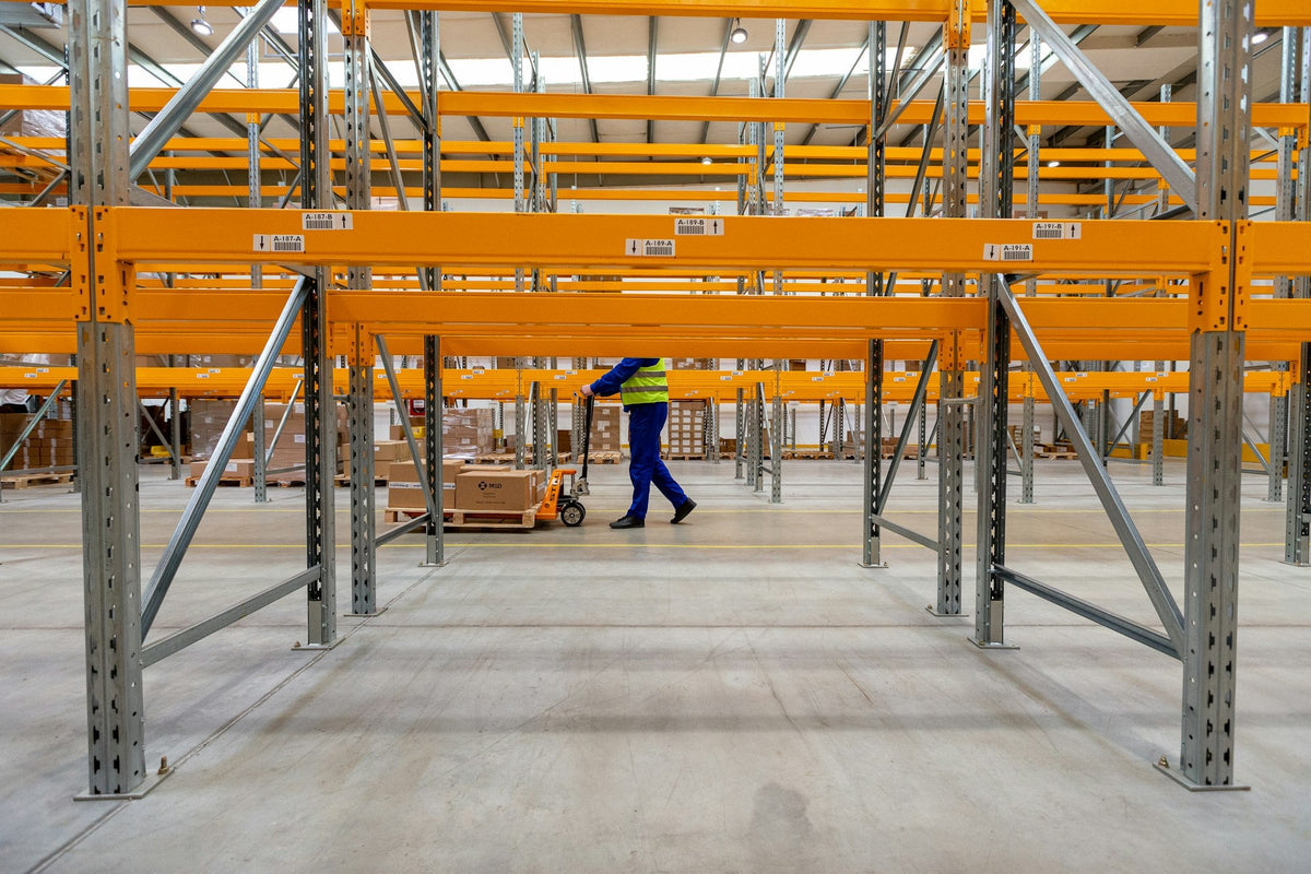 Warehouse worker in a hi-vis vest pulling a loaded pallet jack beneath pallet racking