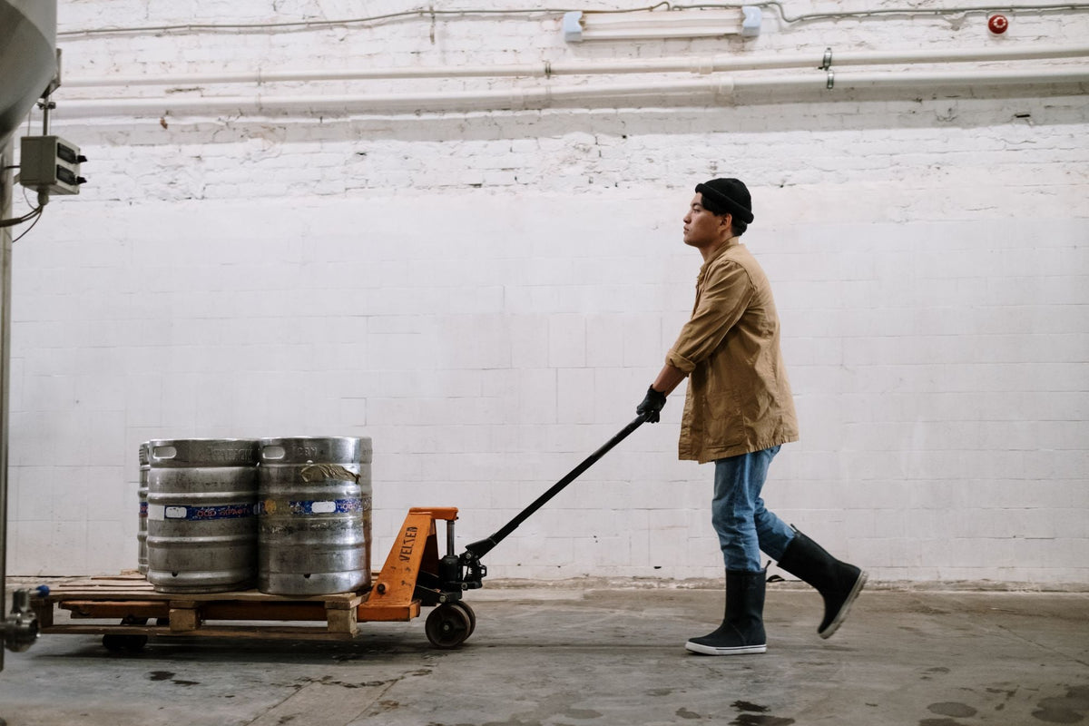 Worker pulling a manual pallet jack loaded with kegs across a warehouse floor